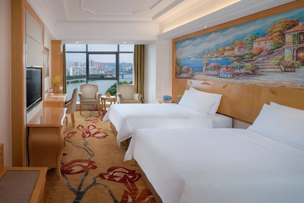 A hotel room with two beds, a TV, a desk, and a painting of a coastal scene.