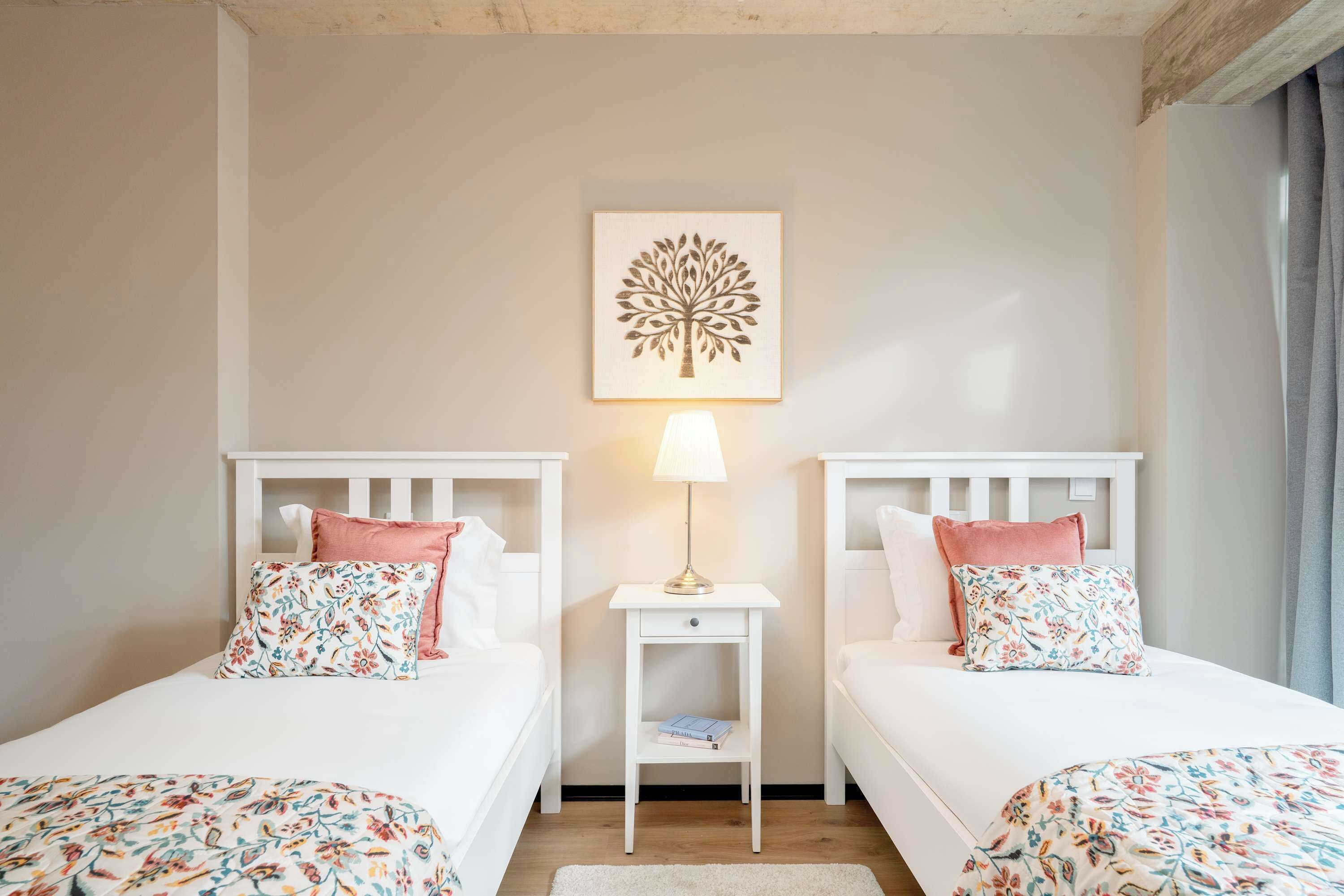 Habitación de hotel con dos camas individuales, cada una con cubrecama y almohadas estampadas con motivos florales. Una mesita de noche con una lámpara y un libro se encuentra entre las camas. Un cuadro enmarcado con un árbol está centrado en la pared sobre las camas.