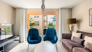 En moderne stue med sofa, to lænestole, sofabord, fjernsyn og en balkon med udsigt over bygninger og grønne områder.