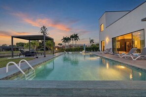 Une piscine avec des chaises longues, un espace de détente couvert et un bâtiment moderne doté de grandes fenêtres.