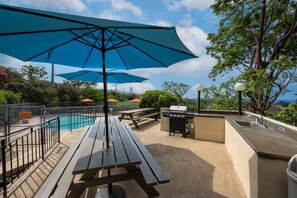 An outdoor pool area with picnic tables, umbrellas, a barbecue station, and a view of the surrounding landscape. - Kona Mansions#b217 Beautiful Unit With Ocean View! (Kailua-Kona)