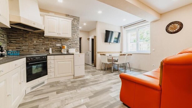 Una cocina moderna con armarios blancos, salpicadero de piedra y horno empotrado. Hay una zona de comedor con mesa y sillas, y un sof á naranja brillante.