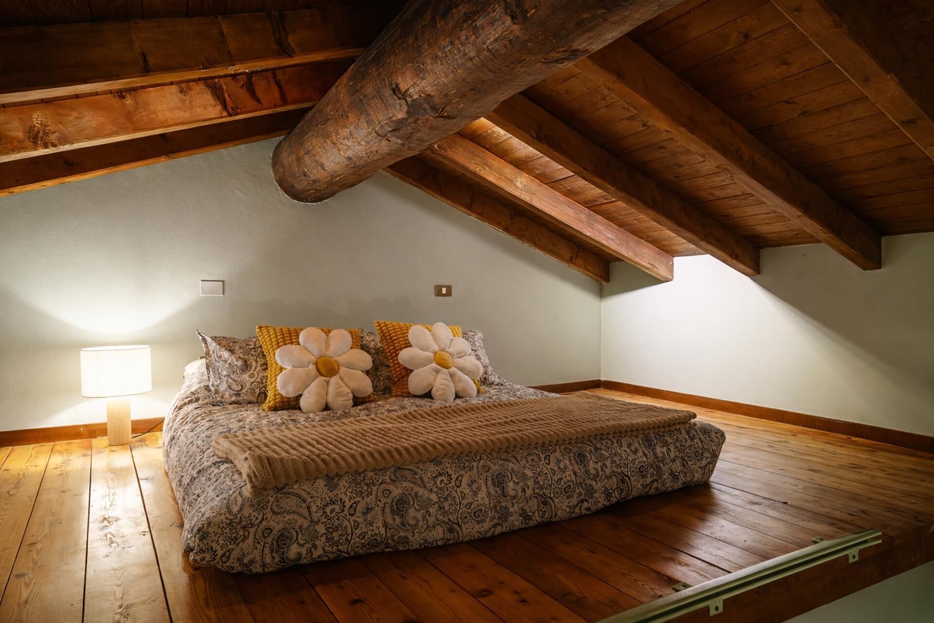 Een slaapkamer met een houten plafond, een bed met bloemenkussens, een bedlampje en een beddengoed met een patroon.
