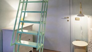 Ein Zimmer mit Holzboden, einer weißen Tür, einer grünen Leiter, einem kleinen weißen Schrank, einer Lampe und einem gemusterten Teppich.