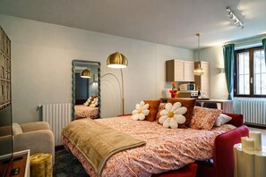 Camera d'albergo con un letto grande, un angolo salotto con una poltrona rossa, una kitchenette e una finestra con tende.