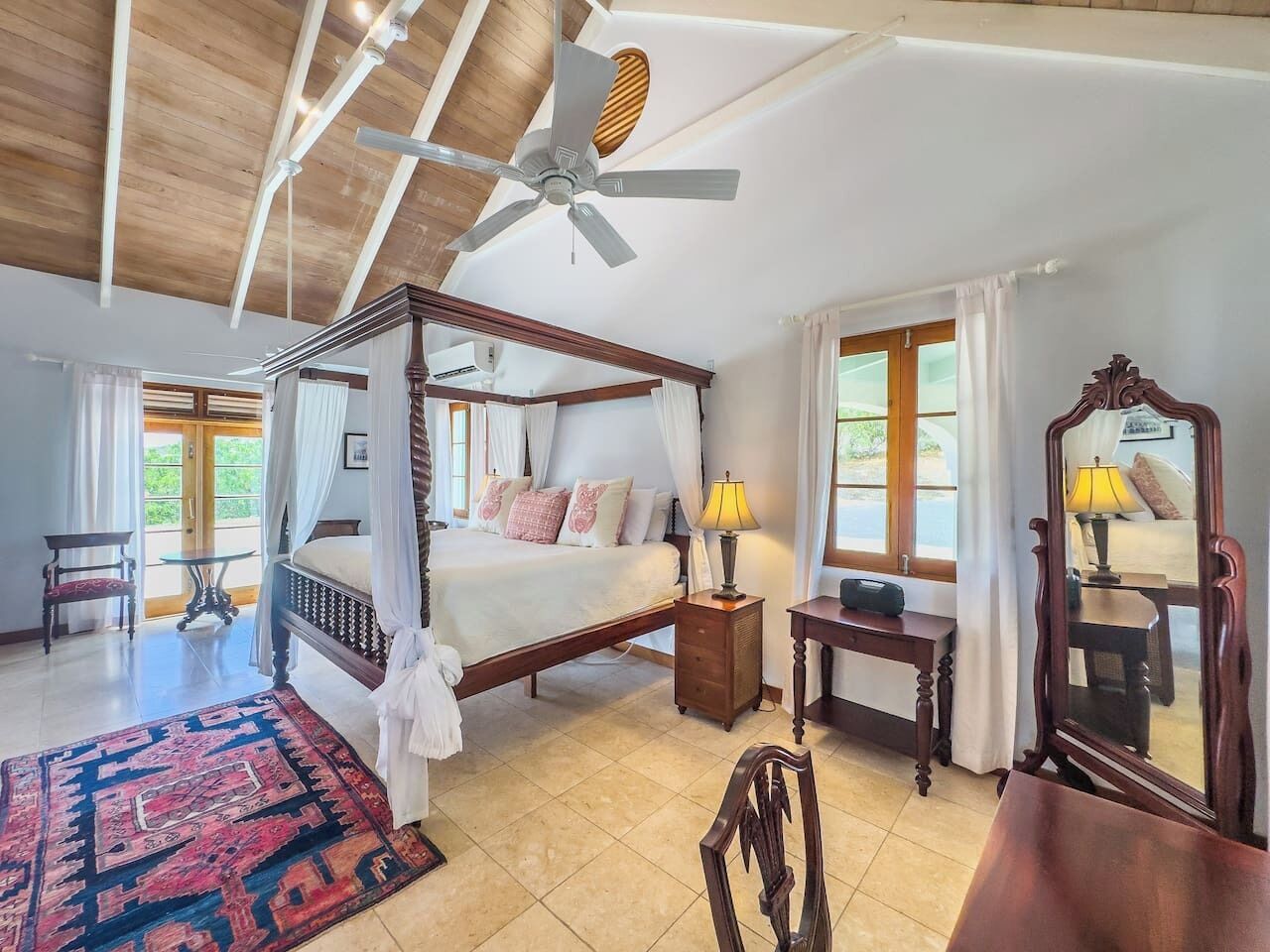 A four-poster bed with a canopy, a ceiling fan, a wooden nightstand, a table with a lamp, and a mirror.