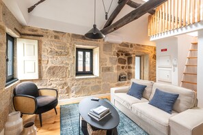 A cozy living room with a stone wall, a white sofa with blue cushions, a black chair, a small table with books, and a hanging light fixture. - Albertina Mezzanine 4 Studio (Porto)