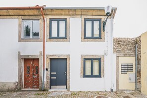 A two-story building with a white facade, a brown door, and a gray door. There are two windows on the upper floor and one on the ground floor. - Albertina Mezzanine 4 Studio (Porto)