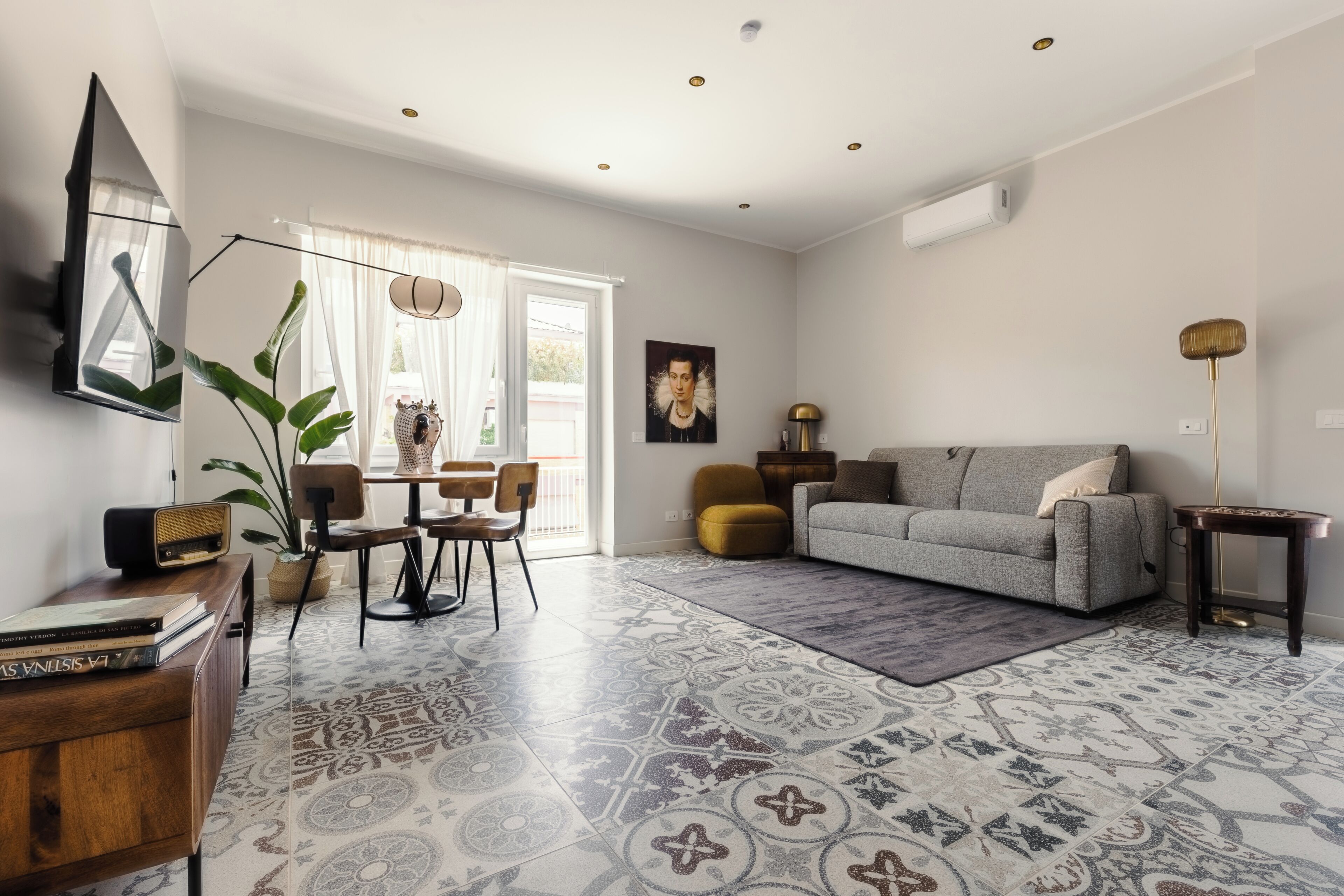 Un salon moderne avec un canapé gris, une table basse, un coin repas avec des chaises, une grande plante en pot et des carreaux de sol à motifs.