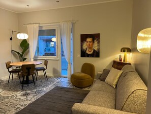 Ein Wohnzimmer mit Sofa, Esstisch mit Stühlen, großem Fenster mit Vorhängen, einem Bild an der Wand und einer Lampe auf einem Beistelltisch.