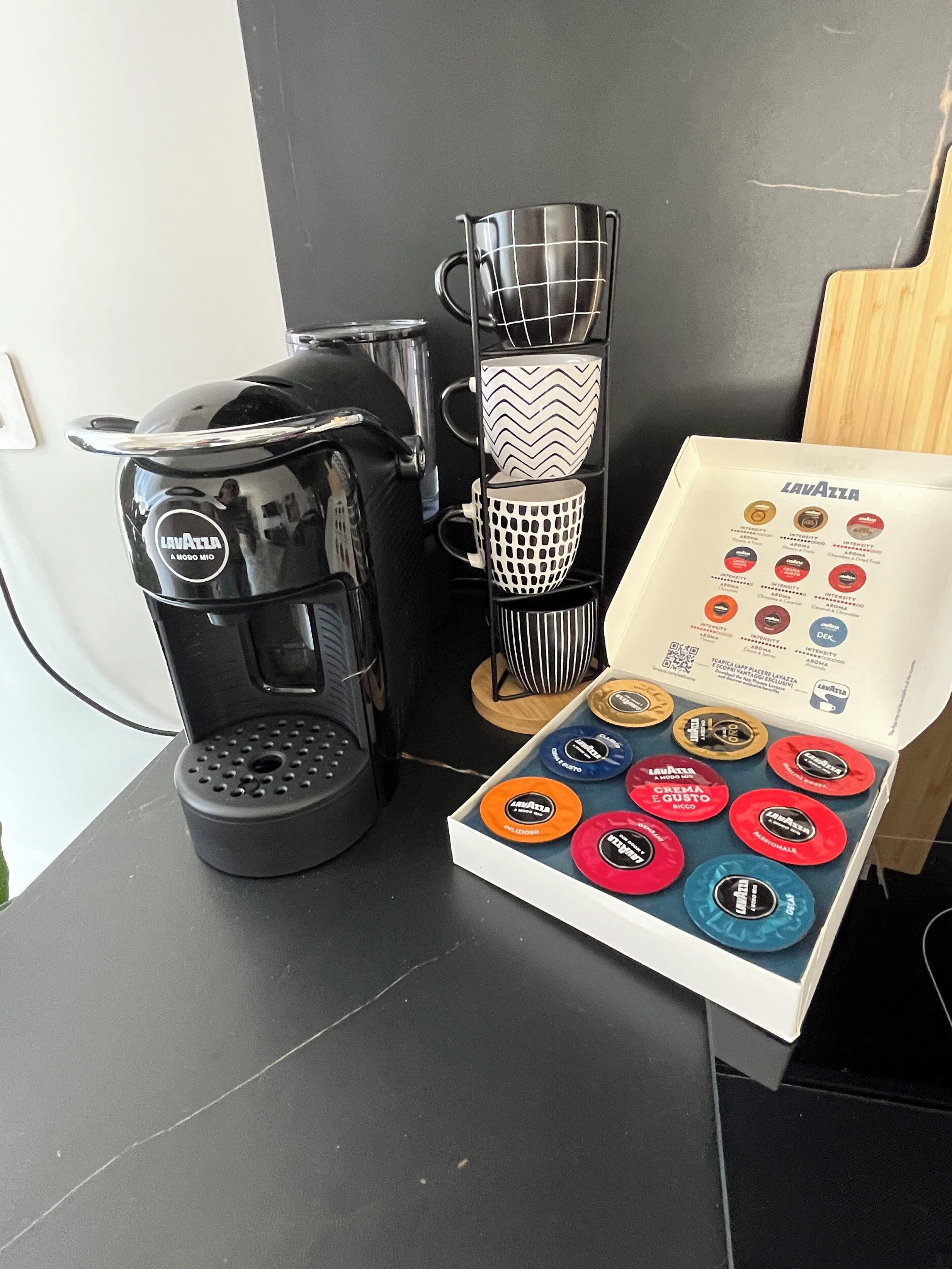 A coffee machine, a set of coffee cups, and a box of coffee capsules on a table.