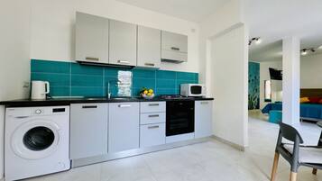 A compact kitchen with a washing machine, microwave, and a small countertop with fruits.