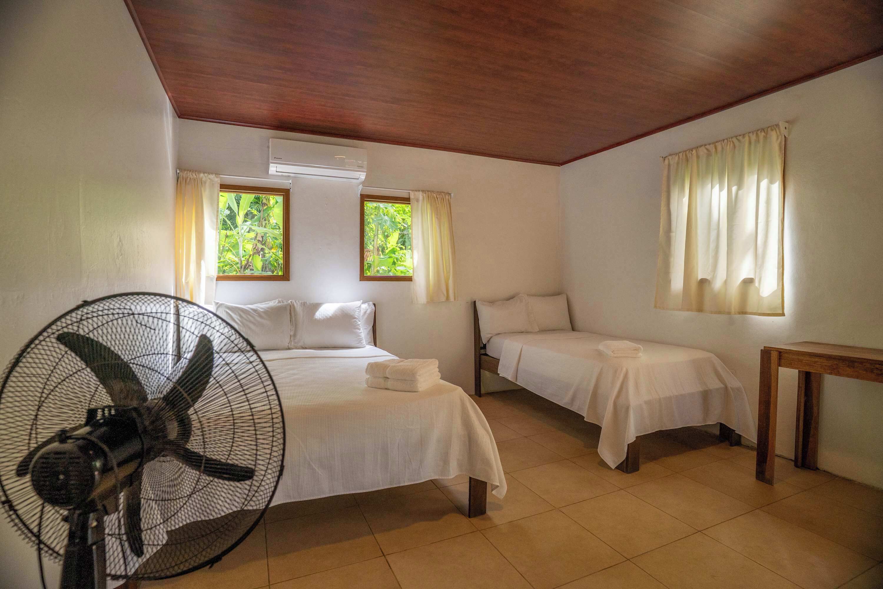 A room with two beds, a standing fan, and a window with curtains.