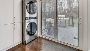 Un cuarto de lavado con lavadora y secadora integradas, una puerta corrediza de vidrio que da a una terraza exterior, y piso de madera.