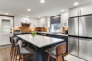 A modern kitchen with white cabinetry, a large stainless steel refrigerator, and a central island with bar stools. - Charming Connecticut Hideaway: Year-Round Fun! (Middlefield)