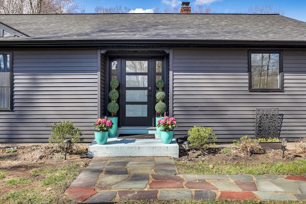 A house with a dark front door, two potted plants with pink flowers, a stone pathway, and a small garden area. - Charming Connecticut Hideaway: Year-Round Fun! (Middlefield)