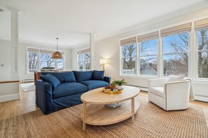 Un salon avec un canapé bleu, un fauteuil blanc, une table basse ronde et de grandes fenêtres équipées de stores.