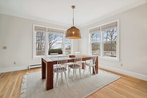 Un espace repas comprenant une table en bois, des chaises blanches, un luminaire suspendu en fibres tressées et de grandes fenêtres équipées de stores blancs.