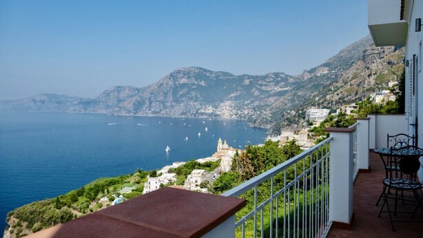 Ein Balkon mit Blick auf das Meer und die Berge, ein weißes Gebäude und Gartenmöbel.