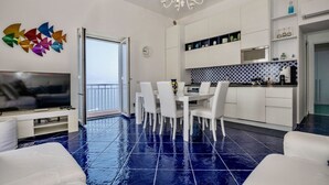 Ein modernes Wohnzimmer mit Flachbildfernseher, ein Essbereich mit weißen Stühlen, eine Küche mit weißen Schränken und ein Balkon mit Blick.