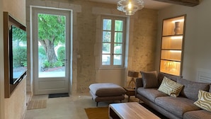 A living room with a sofa, a coffee table, a TV, a lamp, and a window with a view of greenery. - Mas Du Tilleul (Saint-Rémy-de-Provence)