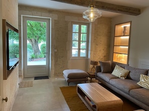 A living room with a sofa, a coffee table, a TV, a lamp, and a window with a view of greenery. - Mas Du Tilleul (Saint-Rémy-de-Provence)