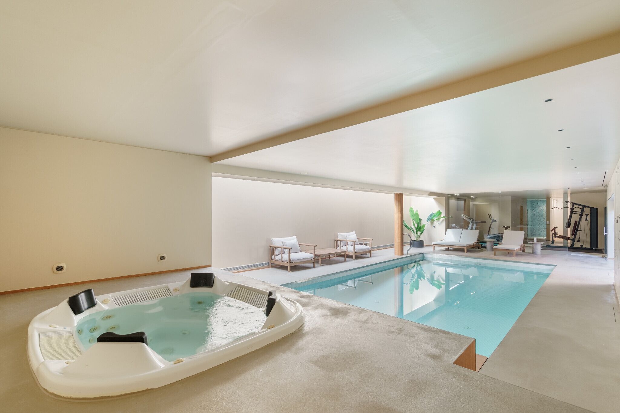 A modern indoor pool with a hot tub, lounge chairs, and exercise equipment.