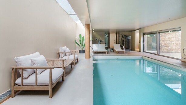Ein moderner Poolbereich im Innenbereich mit freier Sicht auf das Schwimmbecken, eine Sitzecke mit weißen Kissen und einer Topfpflanze.