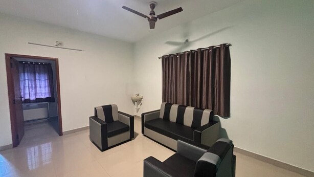 A living room with a sofa set, a coffee table, a side table, a ceiling fan, a window with curtains, and a door leading to another room. - Thamira Residency (Yercaud)
