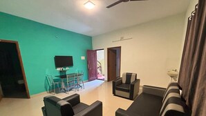 A living room with a teal wall, a flat-screen TV, a dining area with chairs, a sofa, and a ceiling fan. - Thamira Residency (Yercaud)