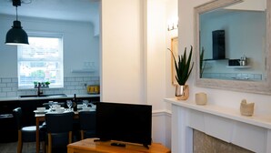 A modern kitchen with a dining area, a TV on a wooden stand, and a fireplace with decorative items. - The Ivies - 5 Bedroom Property - Tenby (Tenby)