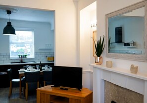 A modern kitchen with a dining area, a TV on a wooden stand, and a fireplace with decorative items. - The Ivies - 5 Bedroom Property - Tenby (Tenby)