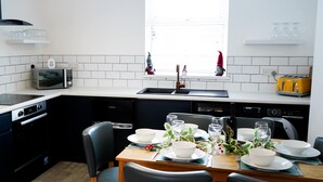 A modern kitchen with white tiles, a microwave, a toaster, and a dining table set for four with plates and glasses. - The Ivies - 5 Bedroom Property - Tenby (Tenby)