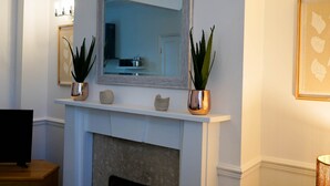 A fireplace with a white mantel, two potted plants, a framed mirror, and a framed artwork on the wall. - The Ivies - 5 Bedroom Property - Tenby (Tenby)