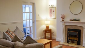 A living room with a sofa, a small table, a lamp, a fireplace, and a door leading outside. - The Ivies - 5 Bedroom Property - Tenby (Tenby)