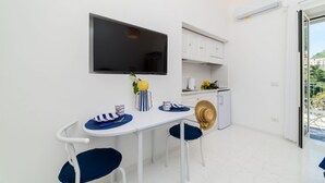 Cozinha compacta com armários brancos, uma televisão de tela plana, uma mesa de jantar com cadeiras e uma geladeira pequena.