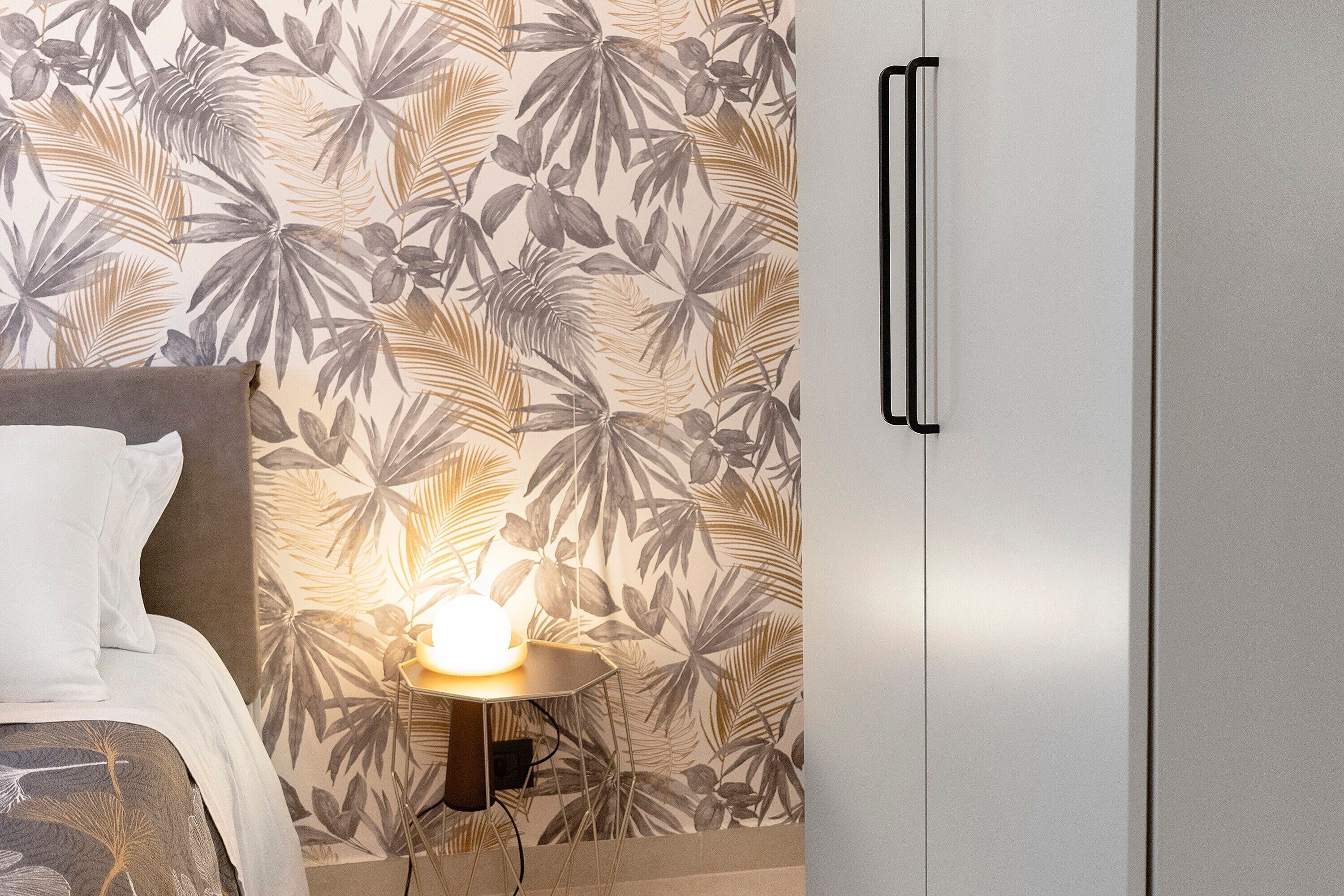 Un dormitorio con cama, una mesita de noche con lámpara, un armario y una pared con estampado de hojas tropicales.