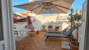 Een terras met een tafel en stoelen, een lounge stoel, potplanten en een grote parasol.