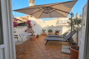 Eine Terrasse mit Tisch und Stühlen, ein Liegestuhl, Kübelpflanzen und ein großer Sonnenschirm.