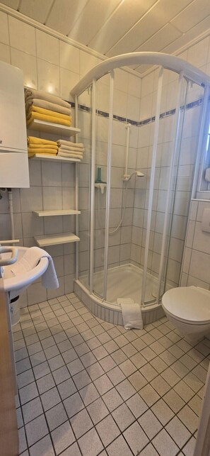Ett badrum med en dusch som är avskärmad med glasväggar, en toalett, ett handfat och hyllor med handdukar.