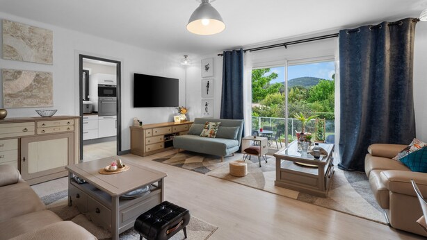 Een moderne woonkamer met een bank, salontafel en een eethoek. Achtergrond toont een keuken met apparatuur en een televisie.