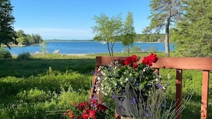 A wooden bench with a flower arrangement in a natural setting with a lake and trees. - Waterfront Home Lake Charlevoix Oyster Bay Mooring Buoy Private Dock Beach fire! (Charlevoix)