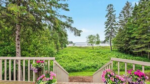 A wooden deck with railings, potted plants, and a view of a green landscape and water body. - Waterfront Home Lake Charlevoix Oyster Bay Mooring Buoy Private Dock Beach fire! (Charlevoix)