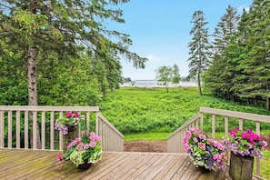 A wooden deck with railings, potted plants, and a view of a green landscape and water body. - Waterfront Home Lake Charlevoix Oyster Bay Mooring Buoy Private Dock Beach fire! (Charlevoix)