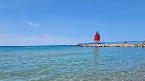 A clear beach with a red lighthouse and rocky shore. - Waterfront Home Lake Charlevoix Oyster Bay Mooring Buoy Private Dock Beach fire! (Charlevoix)