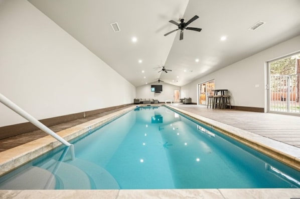Une piscine intérieure dotée de ventilateurs de plafond, d’un téléviseur et d’un espace patio aménagé avec du mobilier d’extérieur.