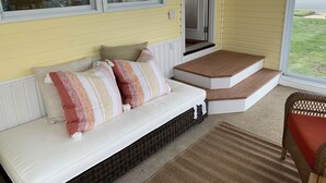 Un porche con un banco blanco, una silla de mimbre y una pequeña escalera de madera que lleva a una puerta.