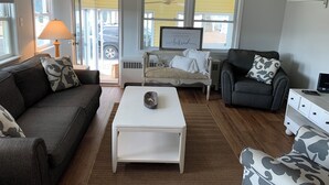 Una sala de estar con sofá, sillón, mesa de centro y televisor. Se ve una ventana con persianas, un ventilador de techo y un aire acondicionado.