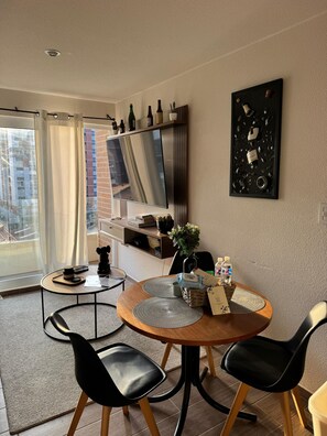 Un espace repas moderne avec une table ronde, des chaises noires, un téléviseur fixé au mur et un balcon offrant une vue sur les bâtiments.