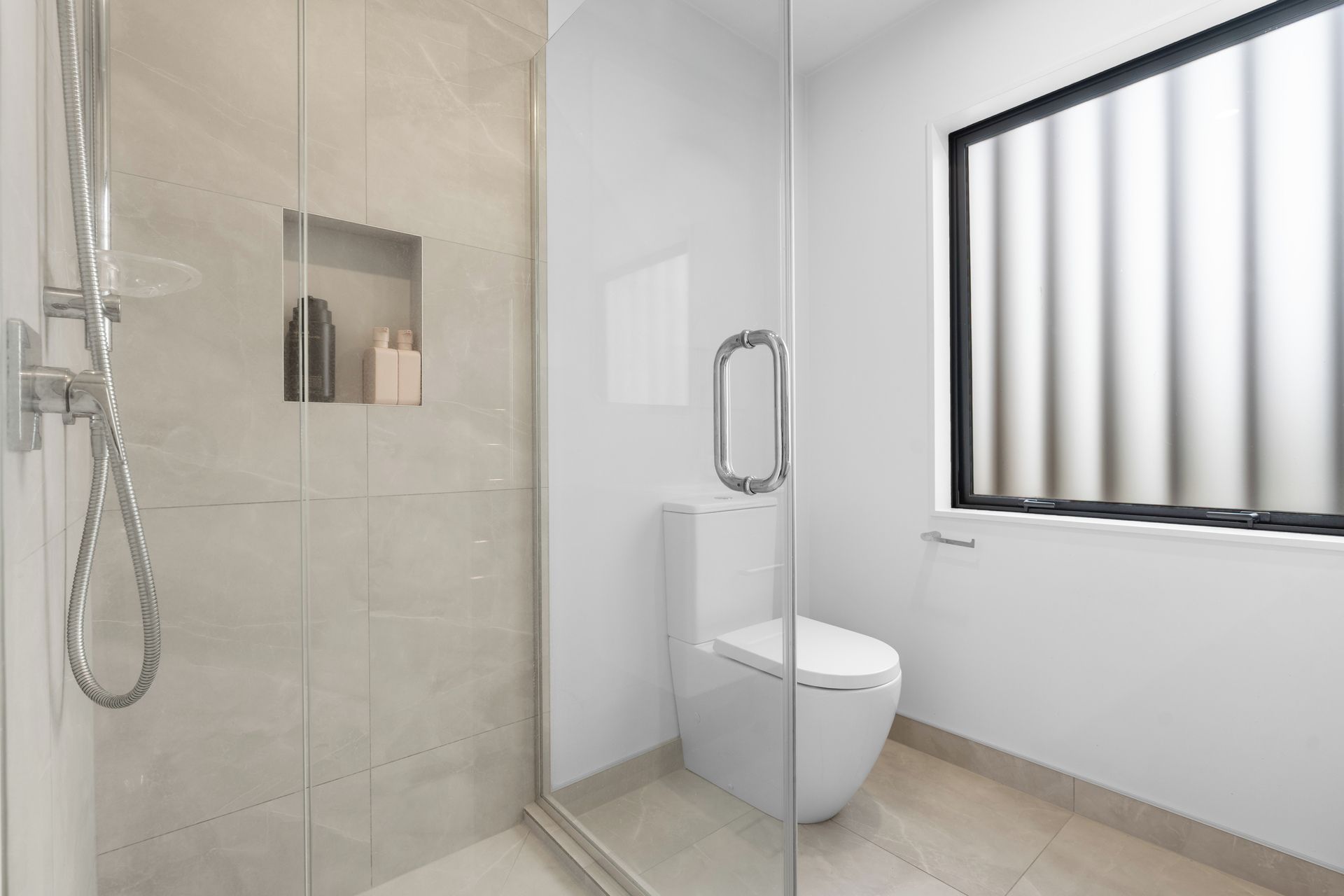 A modern bathroom with a glass shower enclosure, a toilet, and a window with vertical blinds.